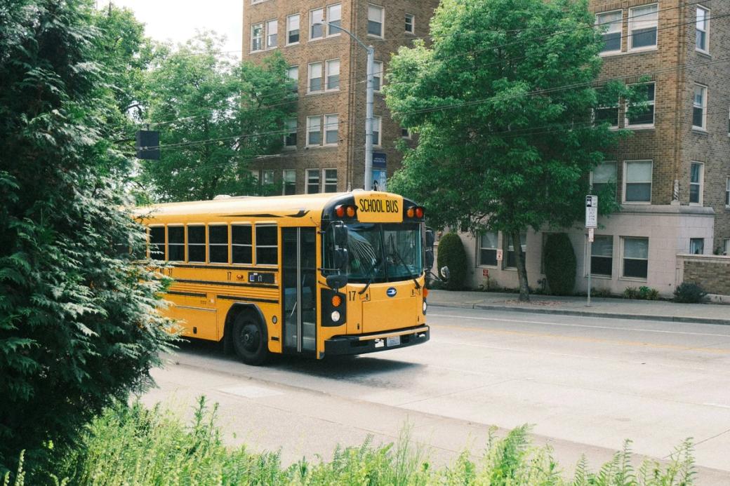 A yellow school bus drives up a hill next to a brick apartment building.
