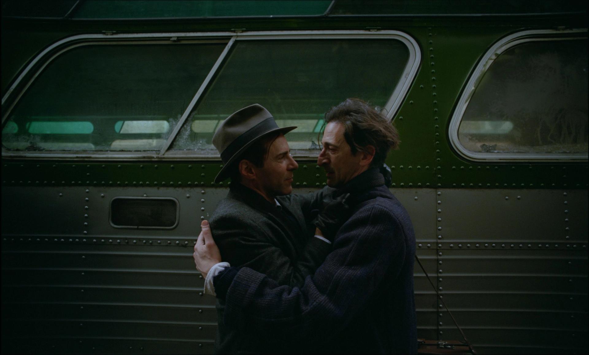 Two men in suits embrace in front of a bus.