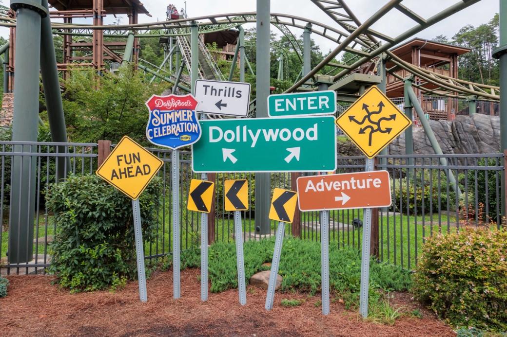 An arrangement of signs at an amusement park. From left to right they read "Fun Ahead" "Summer Celebration" "Thrills" "Dollywood" "Adventure"