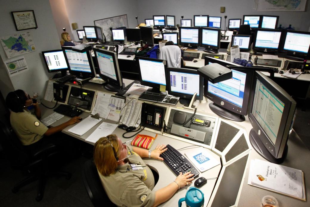 A County Emergency 9-1-1 Dispatch Center in Texas. You can see workers with headsets sitting at desks with multiple monitors preparing to take or actually taking 911 calls.