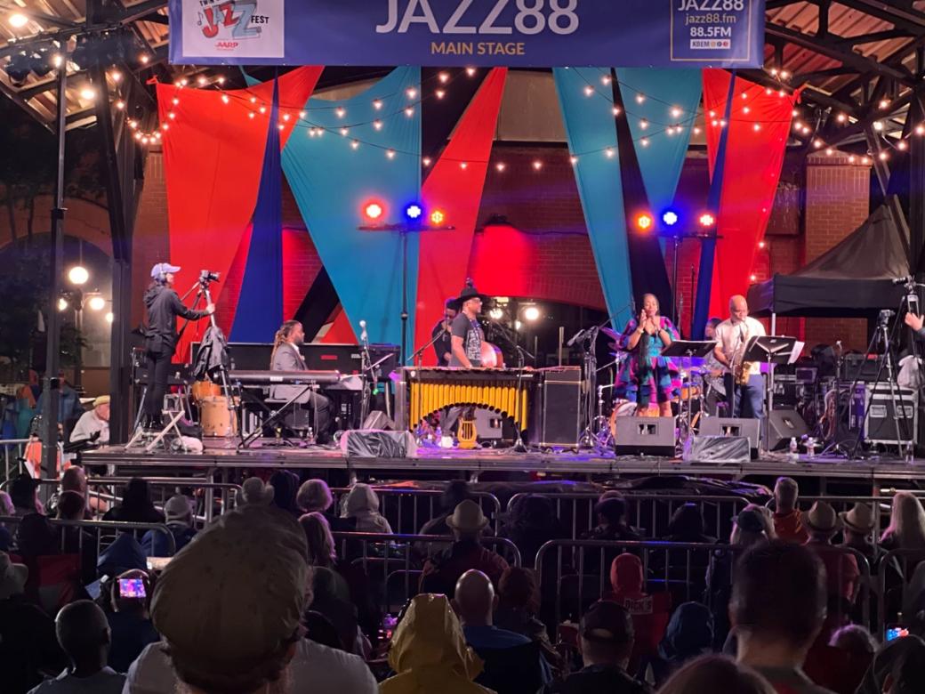 Several musicians on stage, viewed from the audience. Large blue and red pennant flags and a brick wall are behind the musicians.