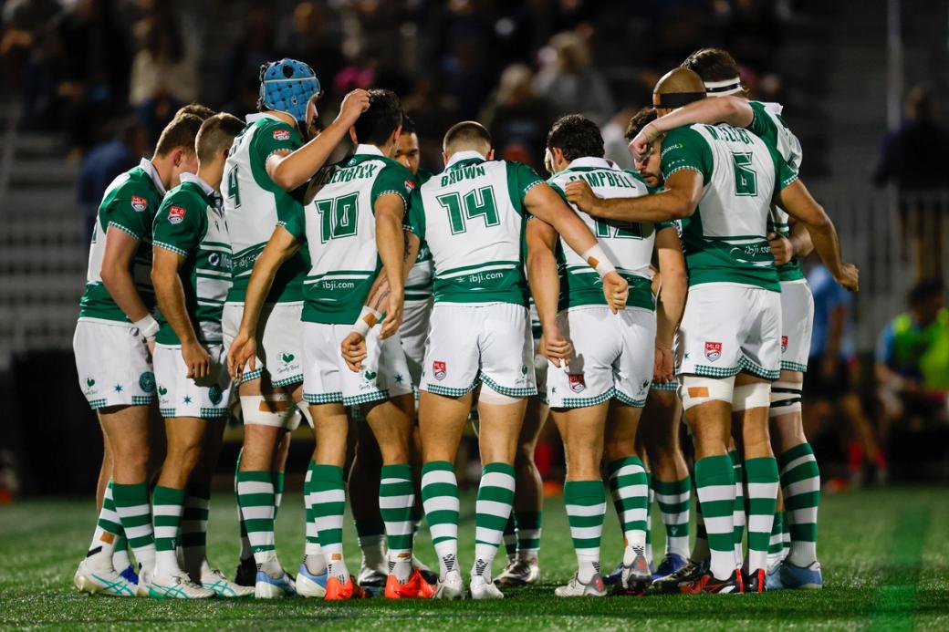 A team of male rugby players wearing green Chicago Hounds jerseys huddle on a green field during a game.