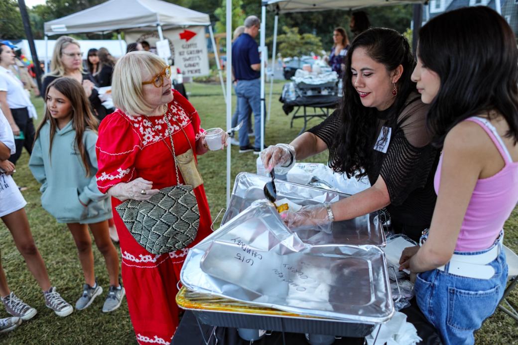 Two women serve food from trays to another woman in a red dress. Other people are in the background.