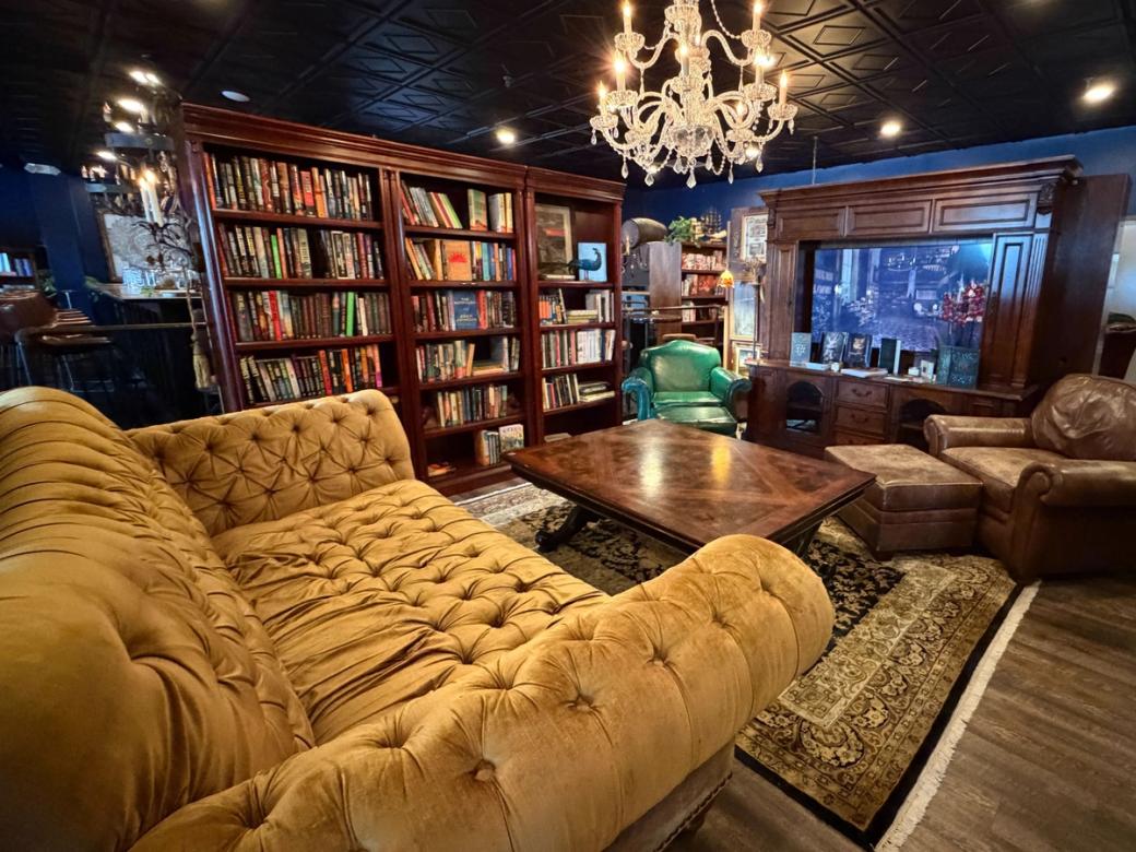 A yellow plush sofa and two leather chairs in front of a wooden coffee table, surrounded by dark wood bookshelves. A chandelier hangs from the black-tiled ceiling.