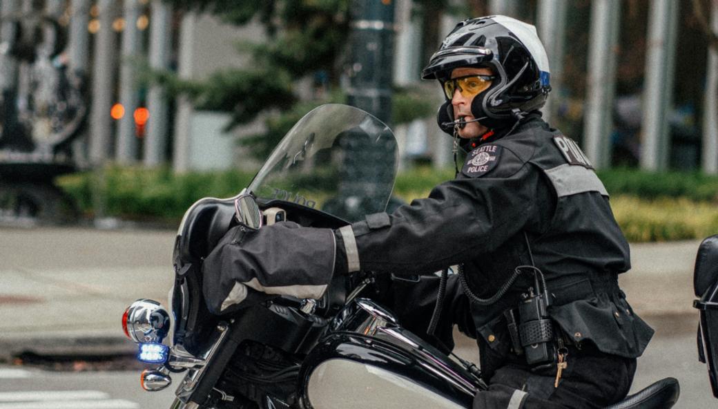 A Seattle police officer sits on a black and white motorcycle at an intersection in front of the downtown library branch. He’s looking to his left through a pair of glasses and a helmet.