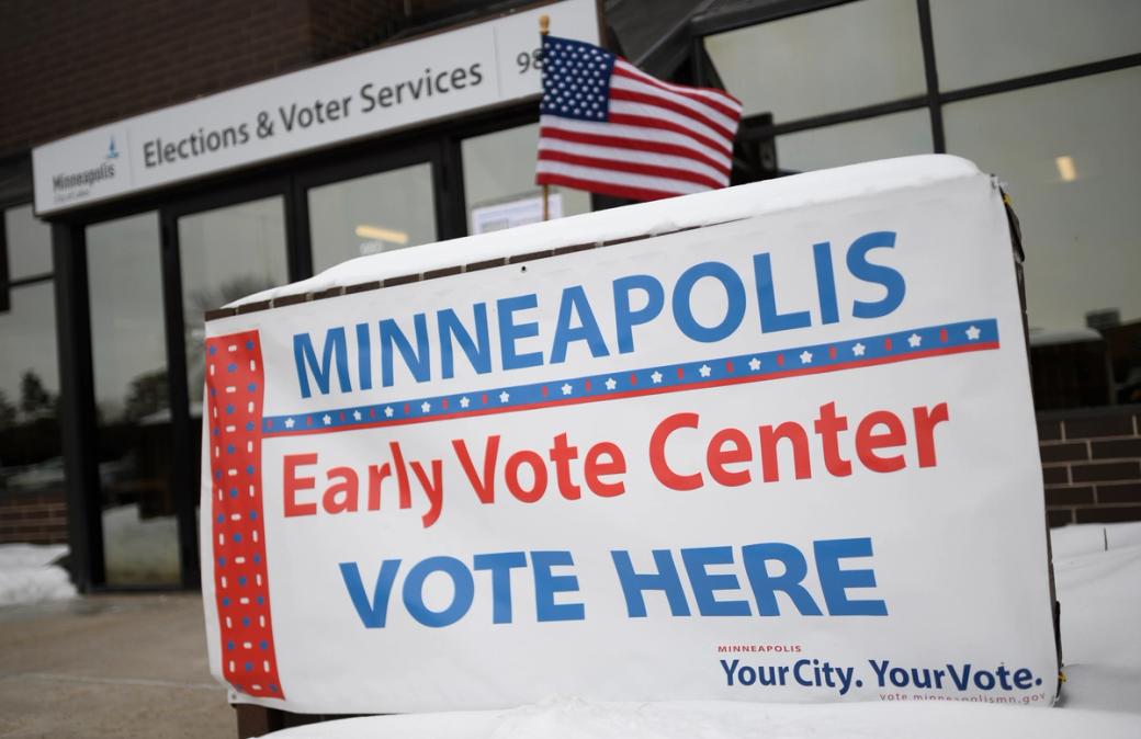 Sign adorned with American flag in foreground that reads, "MINNEAPOLIS Early Vote Center VOTE HERE" and Elections and Voter Services brick building in background