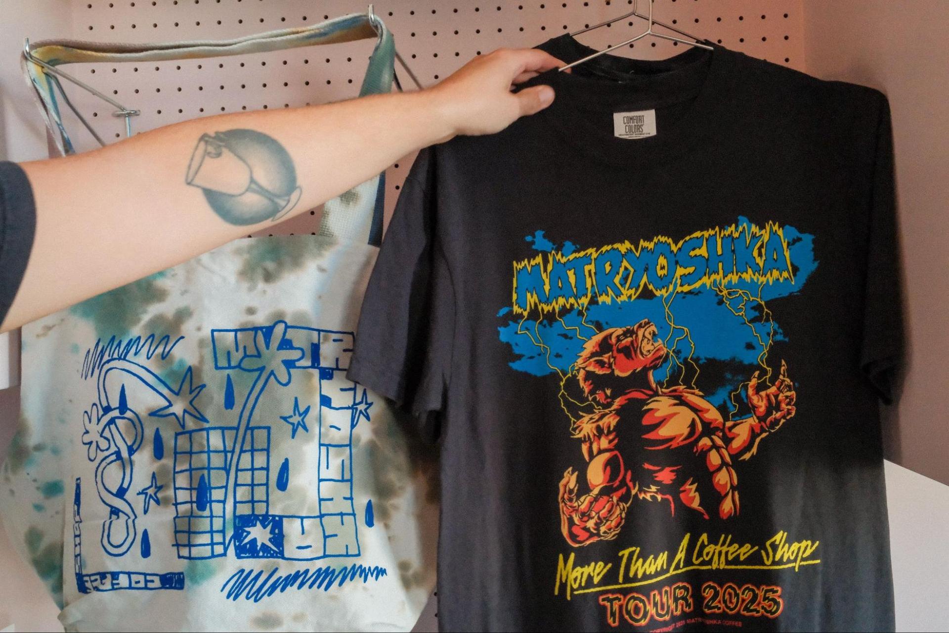 A tie-dyed tote bag hangs to the left of a black T-shirt that reads, “Matryoshka, More than a coffee shop. Tour 2025.”