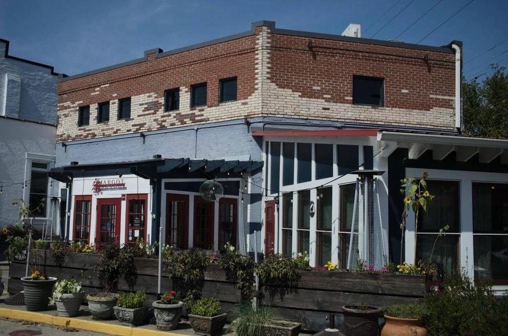 A white and brick restaurant surrounded by planters with flowers.