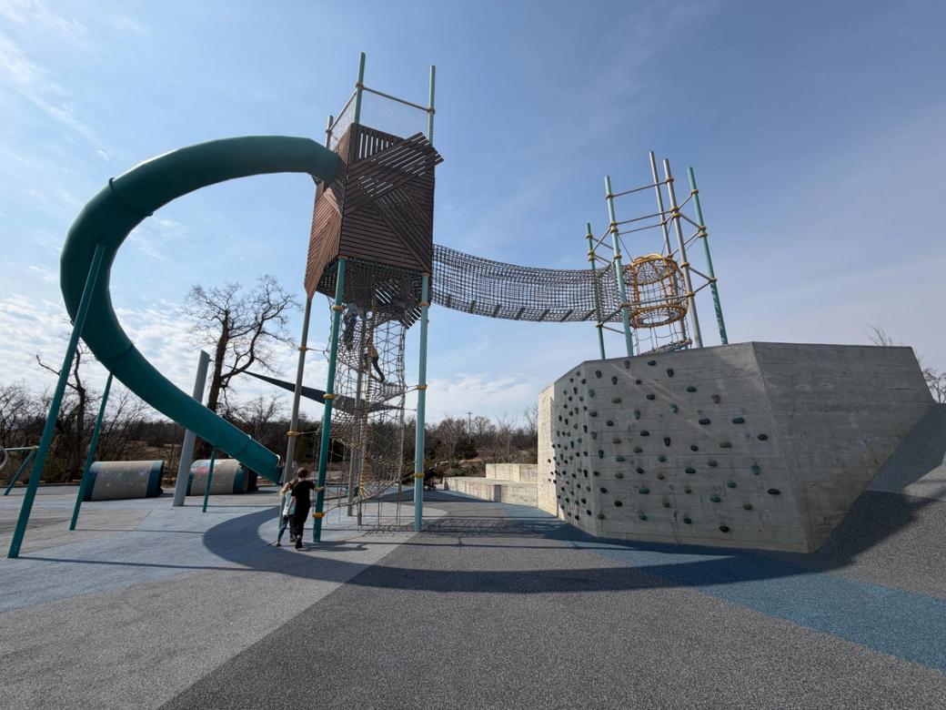 a tall playground structure with climbing ropes leading to a teal slide, beside a climbing rock wall.