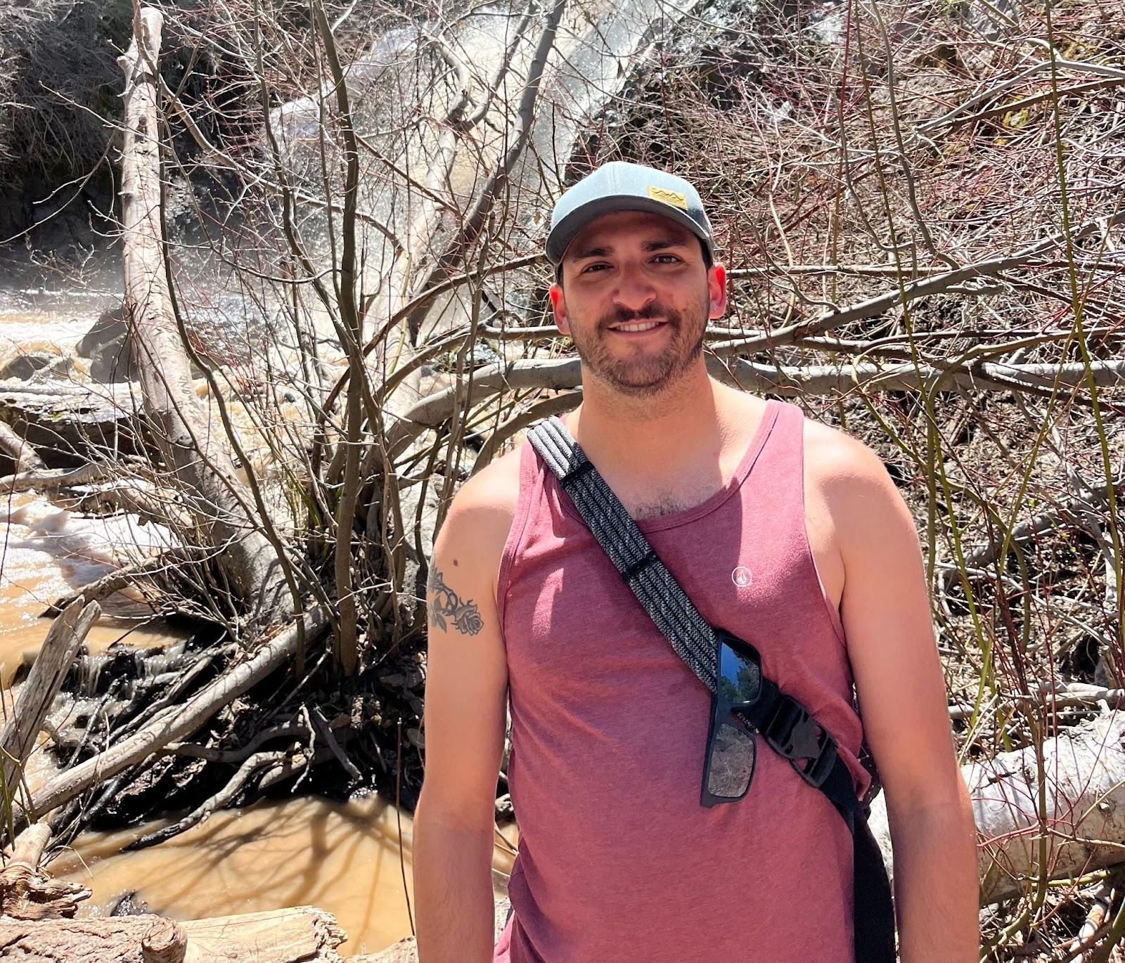 Bearded man in salmon colored tank, black sling bag, and gray hat, stands outdoors in front of a waterfall.