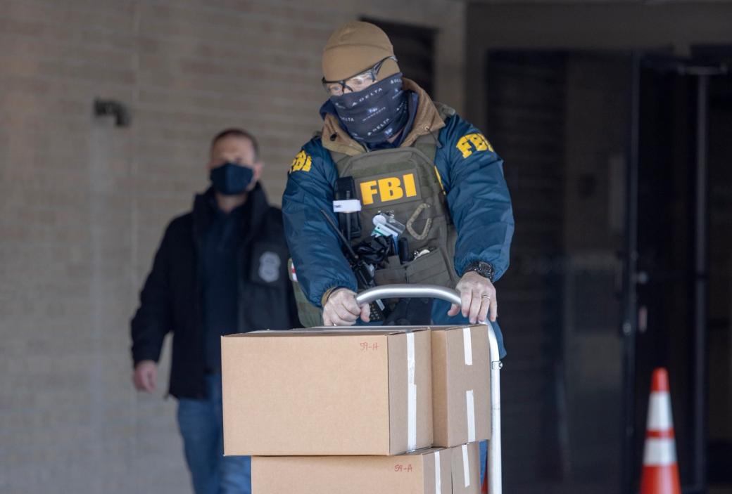 Person in uniform labeled with "FBI" pushes a dolly cart with boxes stacked on it