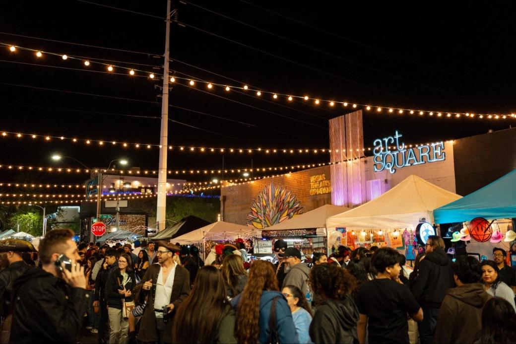 A large crowd of people gathered at night in the art square outside Vegas Theatre Company, with strings of lights above them and booths in the background