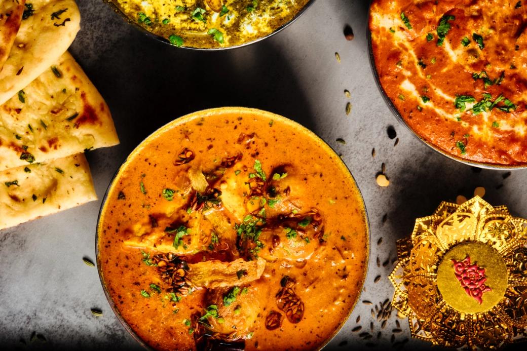 An overhead shot of three bowls of Indian curry — a rich orange-red curry in the foreground garnished with fresh cilantro and whole spices, a green herb-based curry at the top, and a tomato-based dish at upper right — served alongside pieces of garlic naan on a dark stone surface, with a decorative gold ornament in the corner.