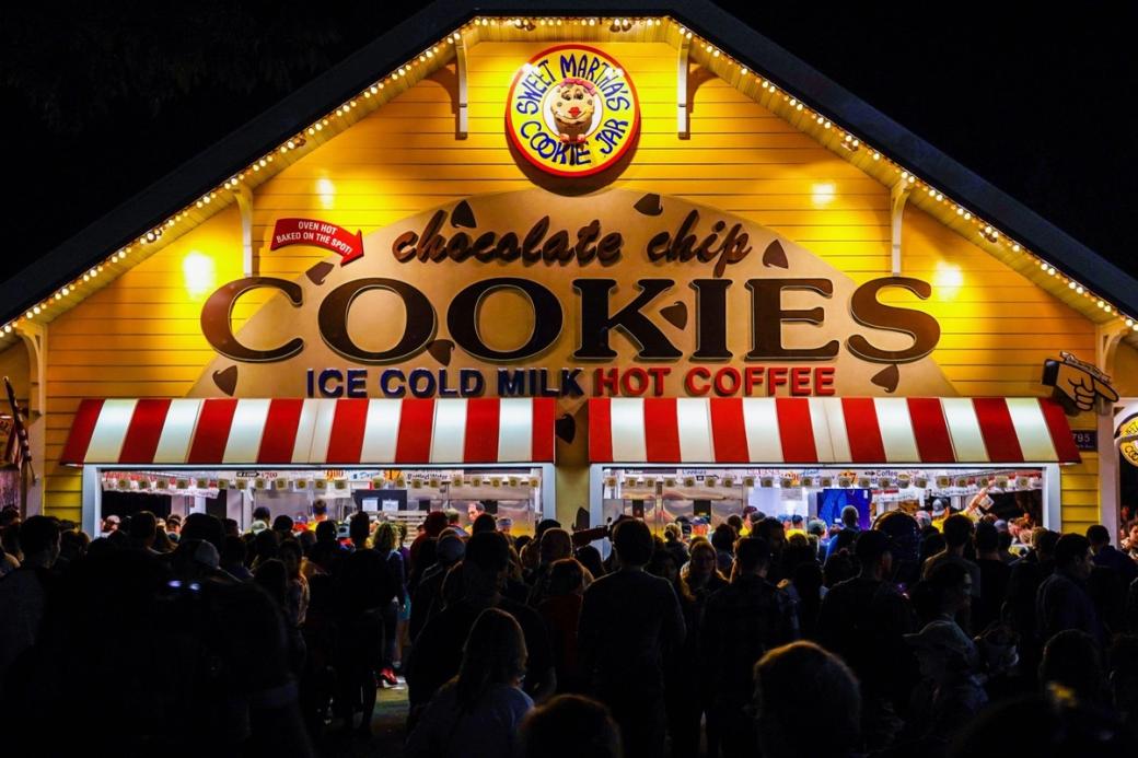 A yellow building with the words “Chocolate Chip Cookies” is illuminated at night as people wait in line.