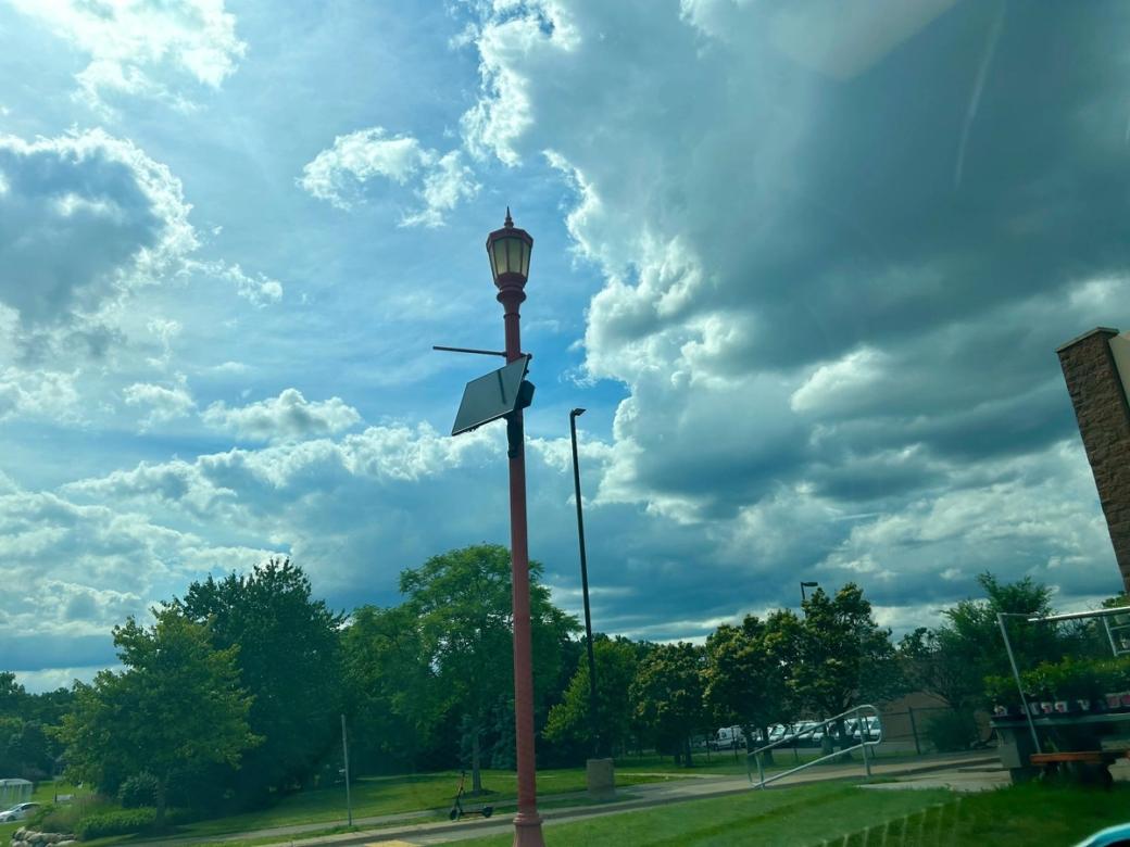 Looking up at lamp post with additional camera attached to it, trees and semi-cloudy skies in the background