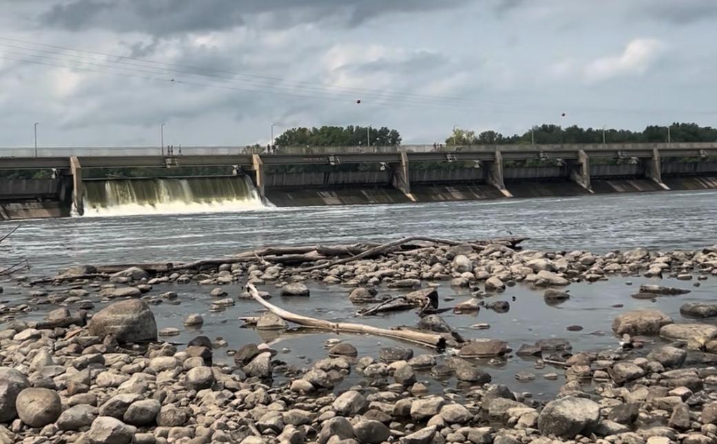 Ground-level view of damn with water flowing and rocky shore in foreground