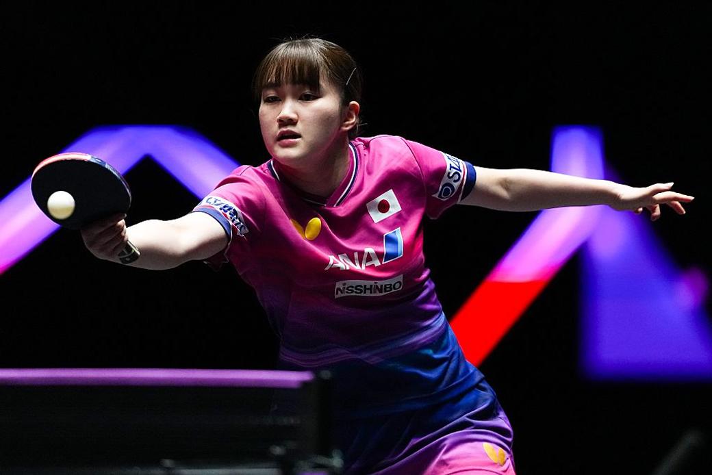 Person hits ball in a table tennis match wearing a pink shirt with purple and black background
