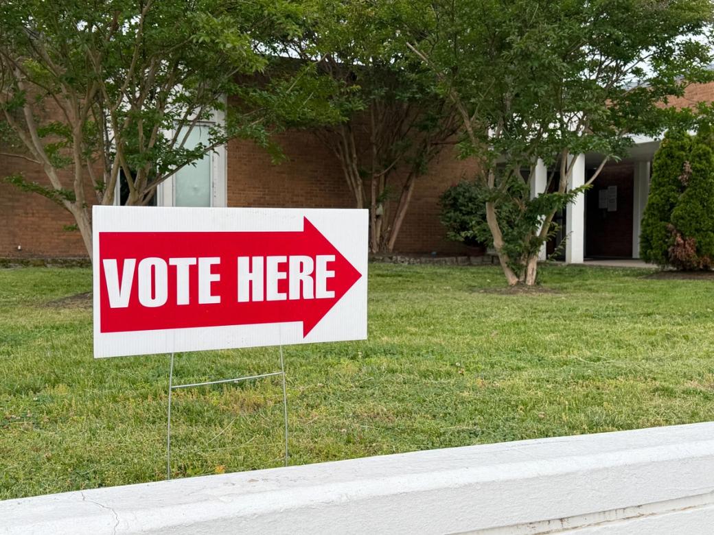 A white voting sign with a red arrow in the grass that says, in white letters, Vote Here. In the background is a brick building and trees.