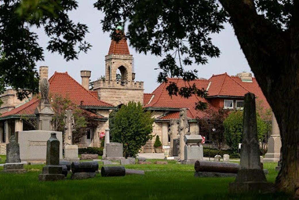 Headstones and monuments in a cemetery in the foreground, with a historic stone building featuring red tile roofs and a bell tower visible among trees in the background.