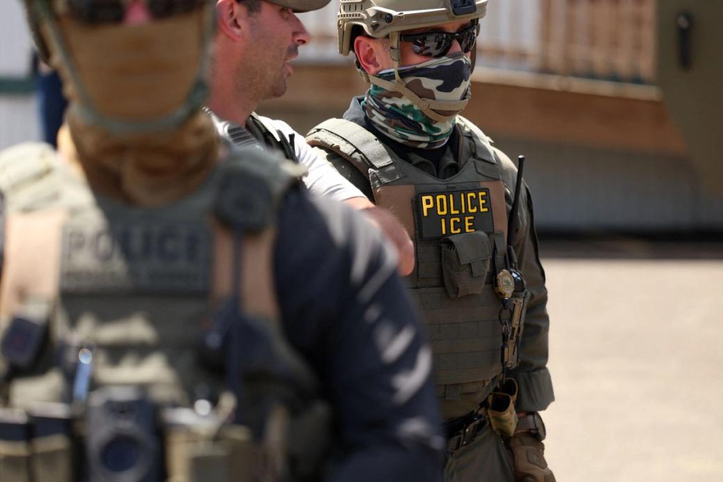 Three ICE agents stand outside a building.