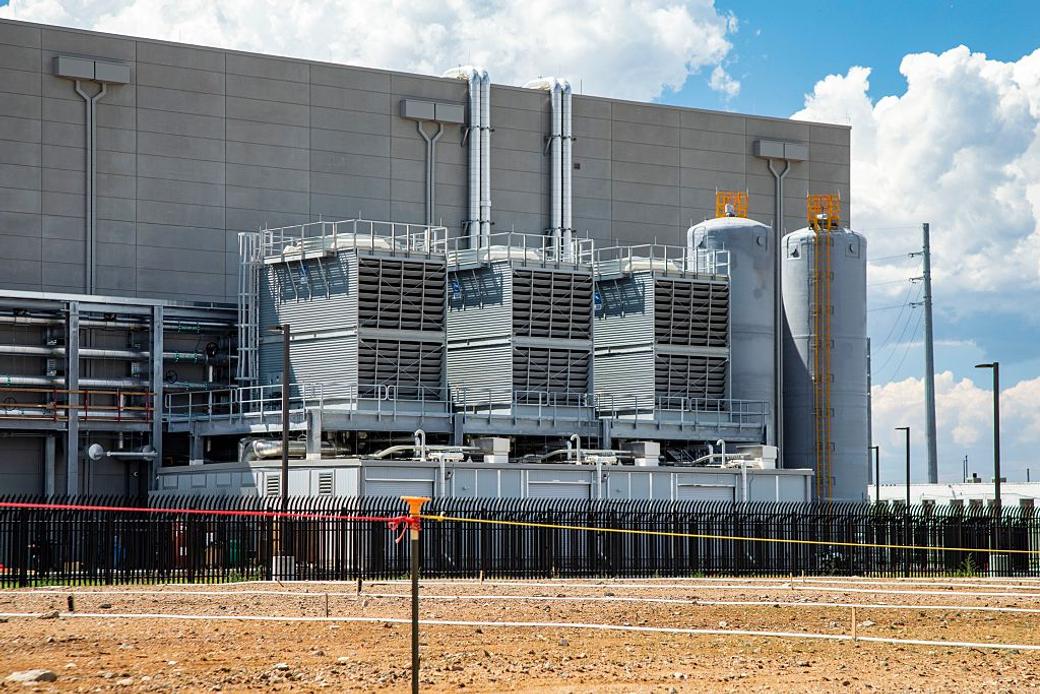 Data center cooling equipment is pictured on the side of a building surrounded by a dirt lot.