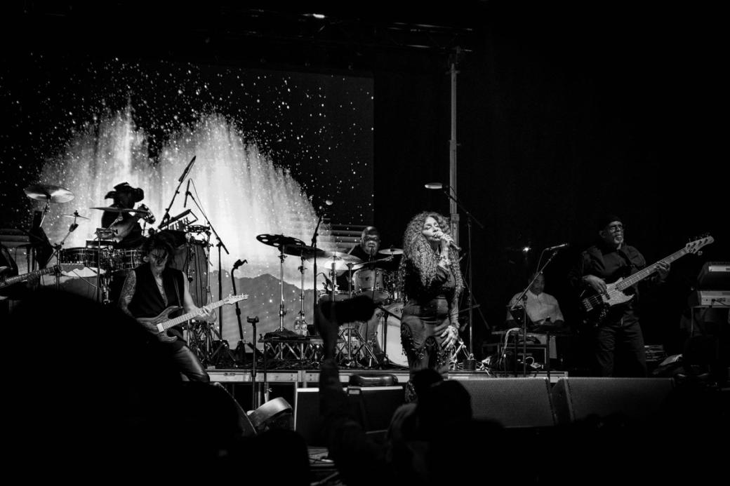 A black and white photo of 5 people on stage. There is one person singing in the front, on the left there is someone playing an assortment of percussion, in the back there is a person on a drumset, on the right there's someone with a bass guitar, off stage there is someone standing