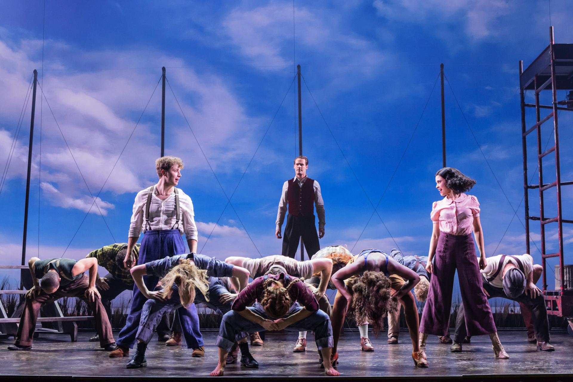 A group of dancers on stage in front of a blue sky backdrop.