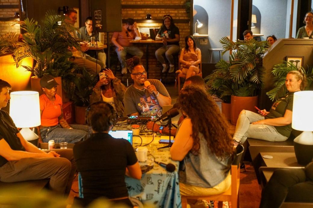 A group of people sitting in a circle with microphones speaking with each other. Around them are people sitting and watching them speak