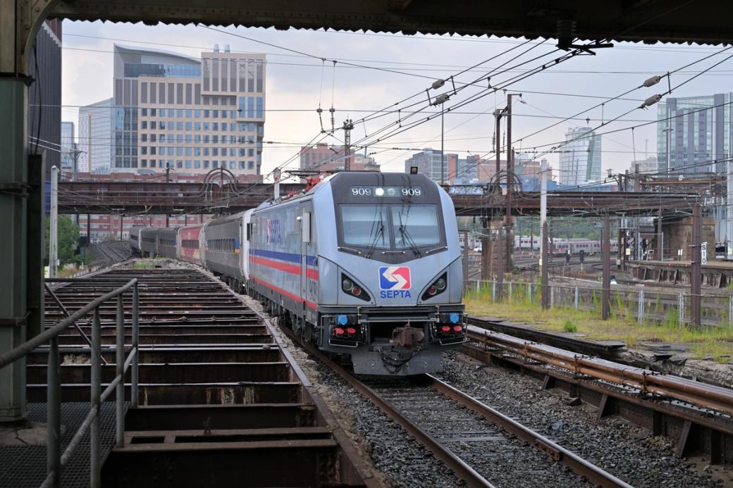 A blue SEPTA locomotive pulls train cars into 30th Street Station. Wires hang above the train cars as it enters the tunnel. Parts of West Philly’s skyline are visible in the background. 