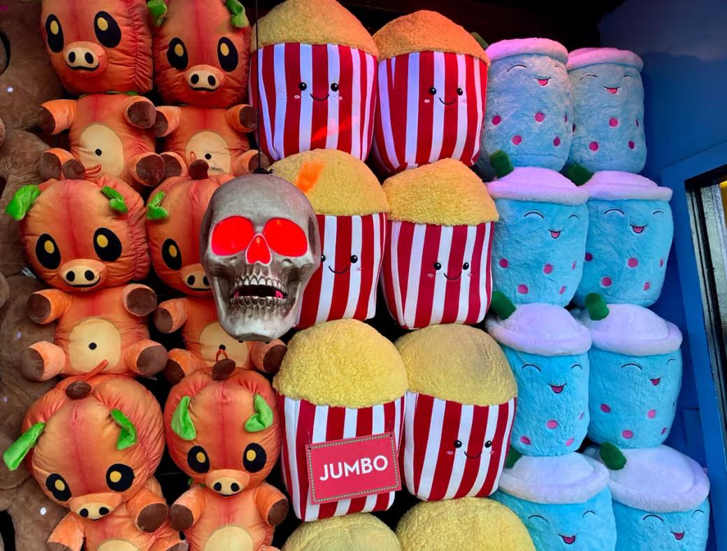 A display packed with oversized plush prizes, including orange pig-like characters with pumpkin heads, smiling red-and-white popcorn buckets, and blue drinks, with a glowing red-eye skull hanging in the center.