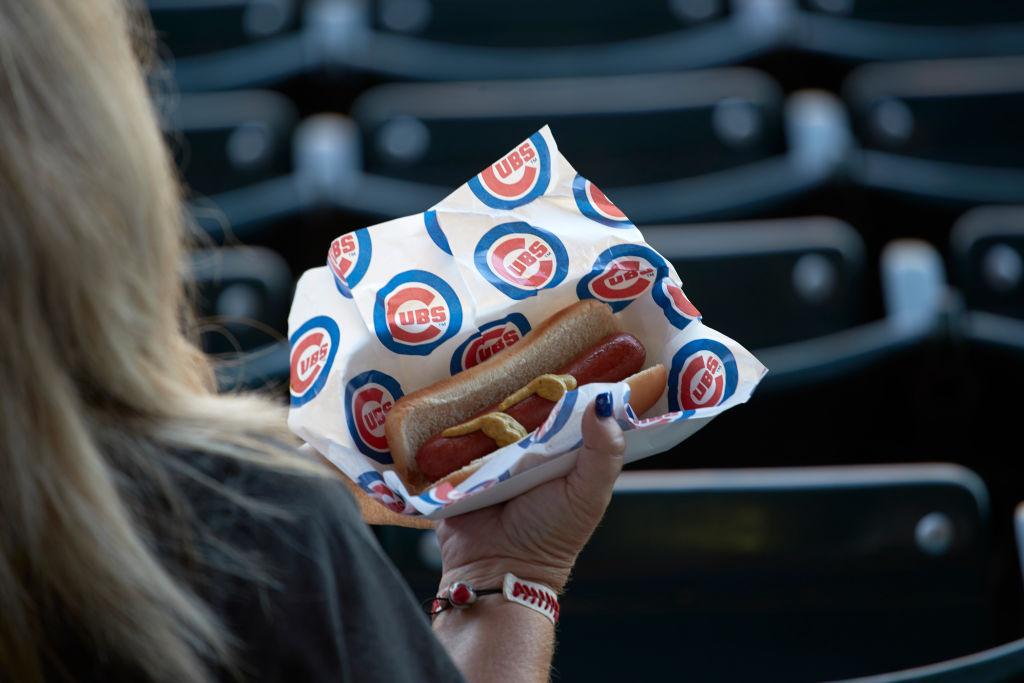 A blonde woman holds a tray in her right hand containing a hot dog with mustard wrapped in Cubs paper