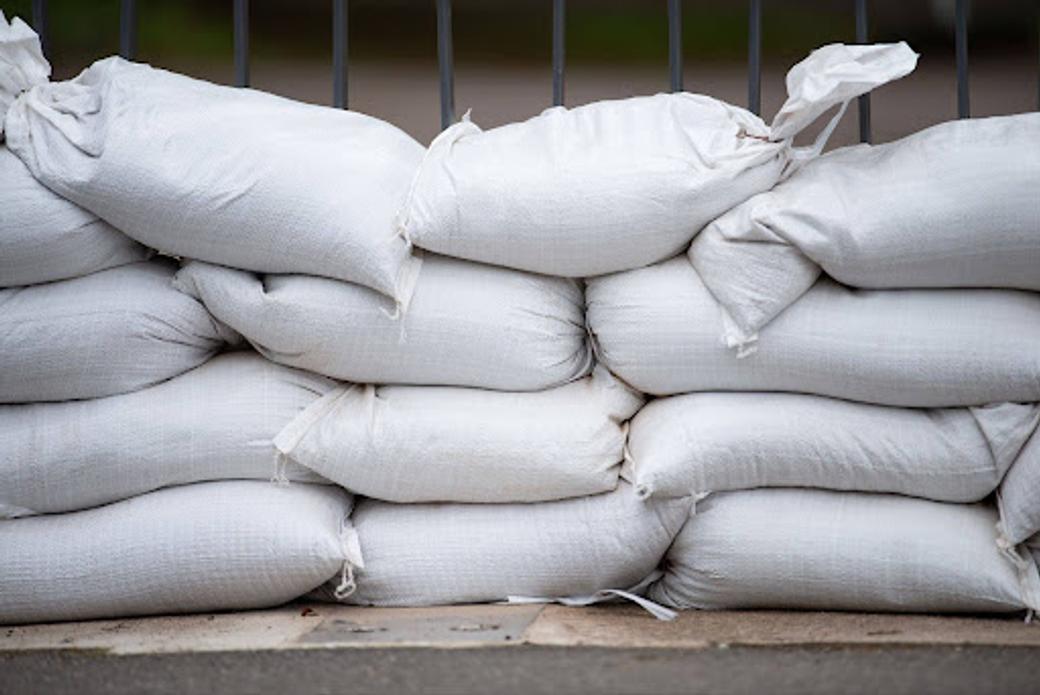 A wall of white sandbags stacked four rows high along a sidewalk with a black metal fence behind.