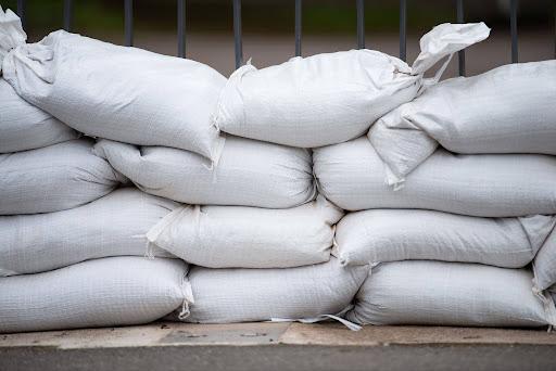 A wall of white sandbags stacked four rows high along a sidewalk with a black metal fence behind.