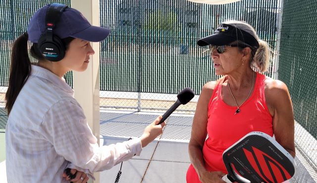 Image of Sonja wearing headphones holding up a mic to interview Patti, a pickleball player in a bright orange tank top, holding a paddle.