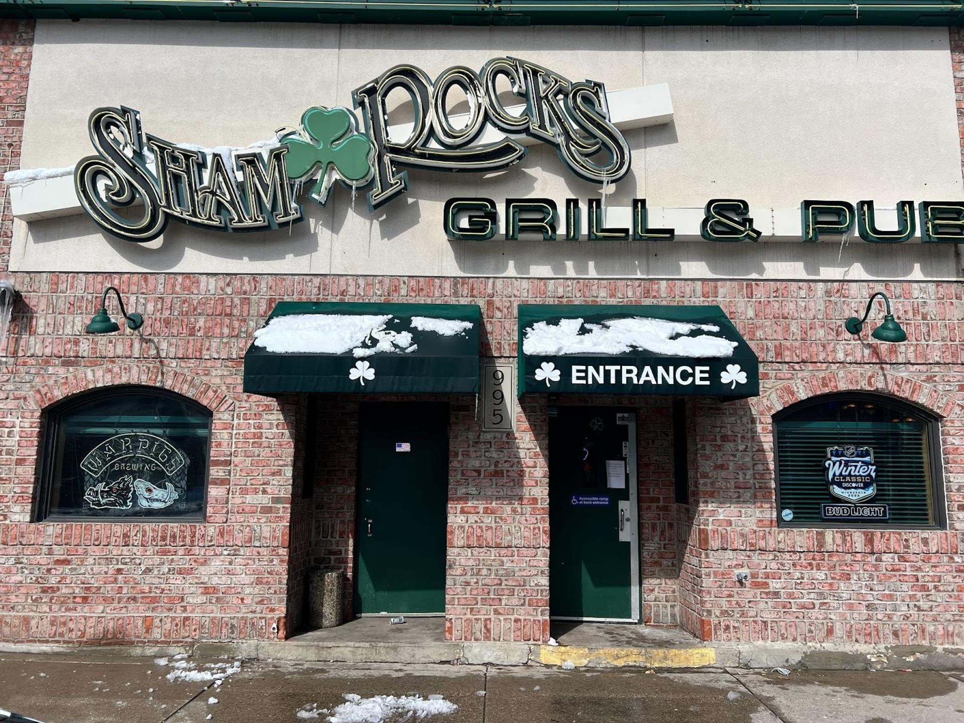 A faded brick building. A neon sign reads "Sham Rocks Grill & Pub" There is a clover between "Sham" and "Rocks"