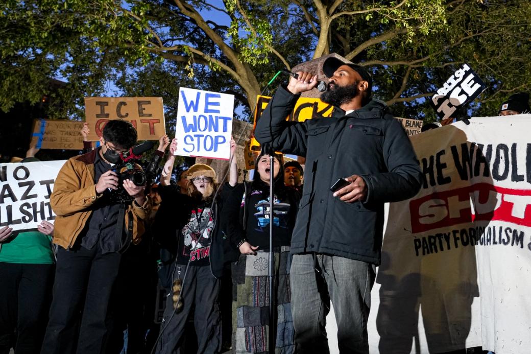 A man wearing a jacket holds a microphone and speaks in front of protesters holding signs.
