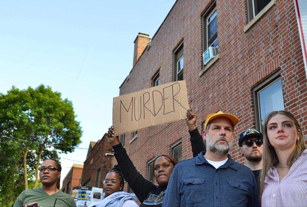 A group of protestors gather on the street with a sign reading "MURDER"