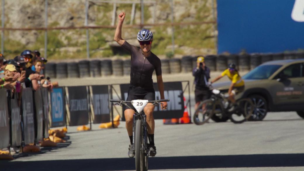 A cyclist on a bike with her right fist in the air. She's wearing a helmet and sunglasses.