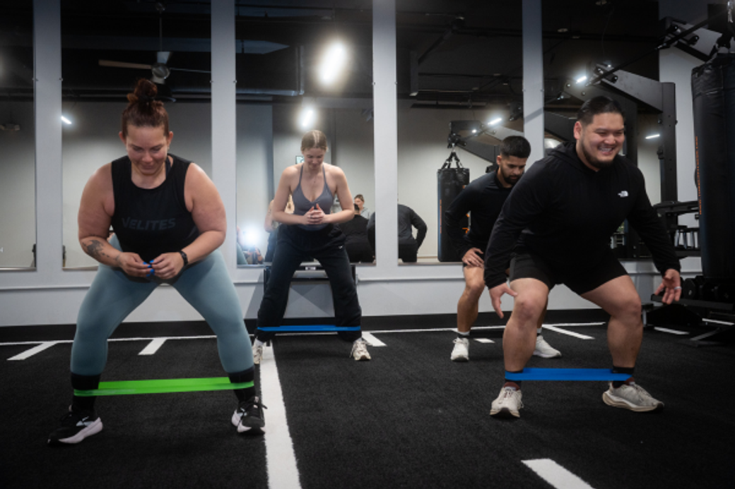 Four people exercise with resistance bands in a gym, smiling as they squat. The atmosphere is energetic, with a focus on fitness and teamwork.