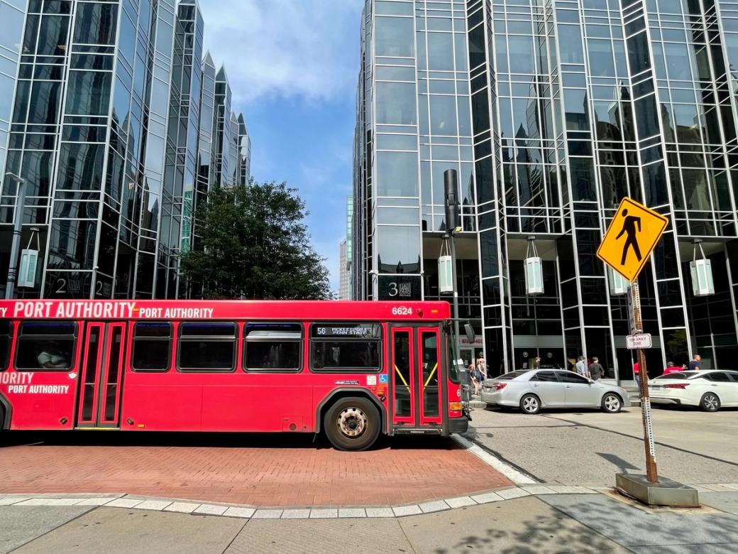A red city bus drives on a street surrounded by other cars, tall reflective buildings, and a pedestrian crossing sign.