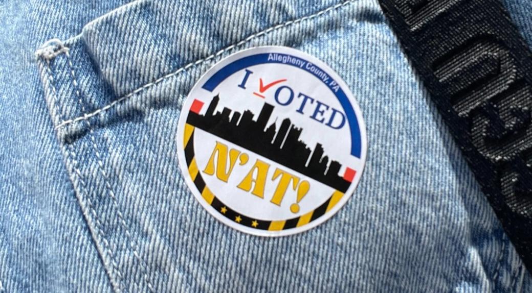 A round sticker that reads “I VOTED N’AT!” with a silhouette of the Pittsburgh skyline is stuck to a denim pocket.