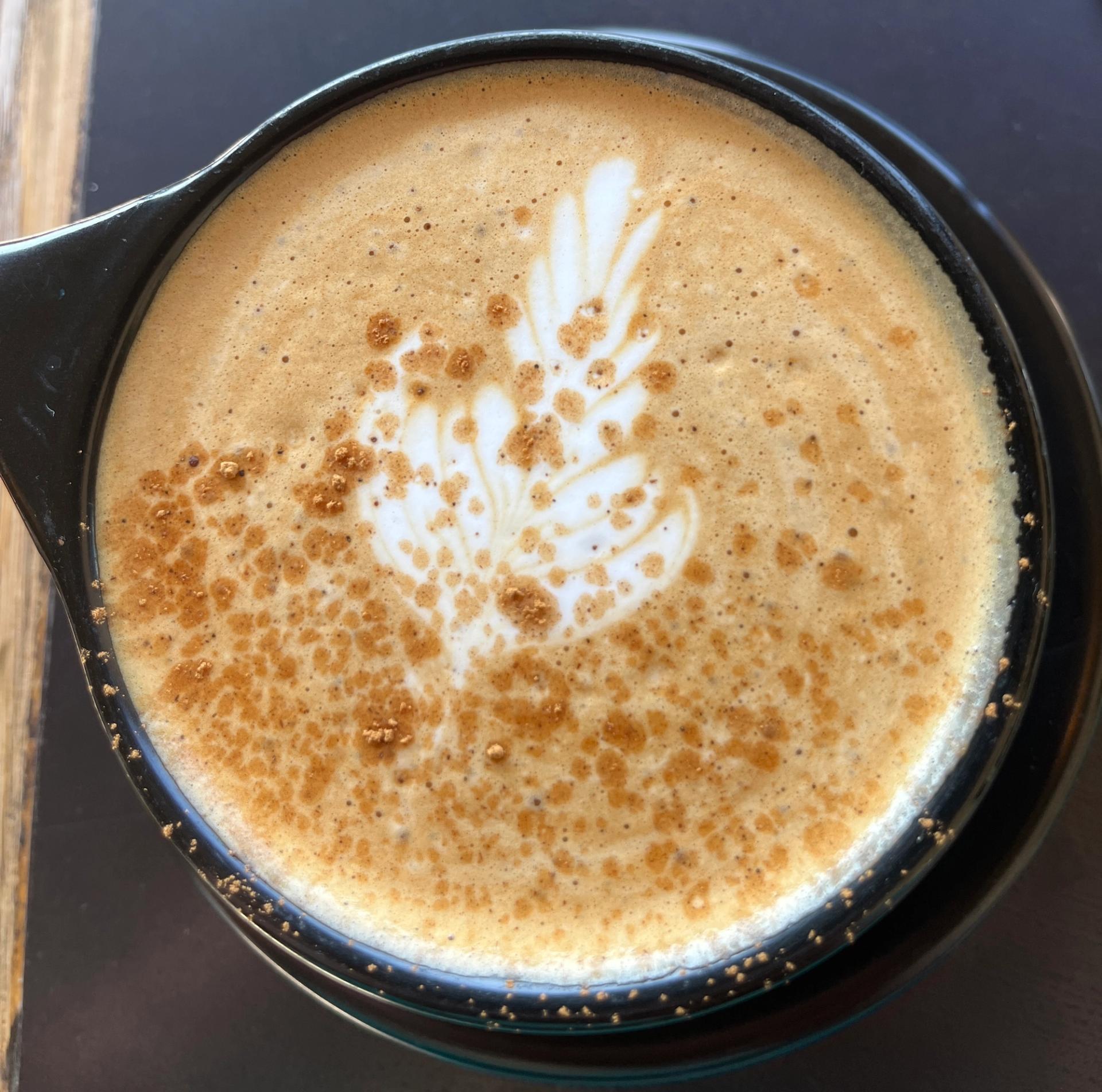An overhead view of a frothy latte with a floral milk art design dusted with spice, served in a black ceramic mug.