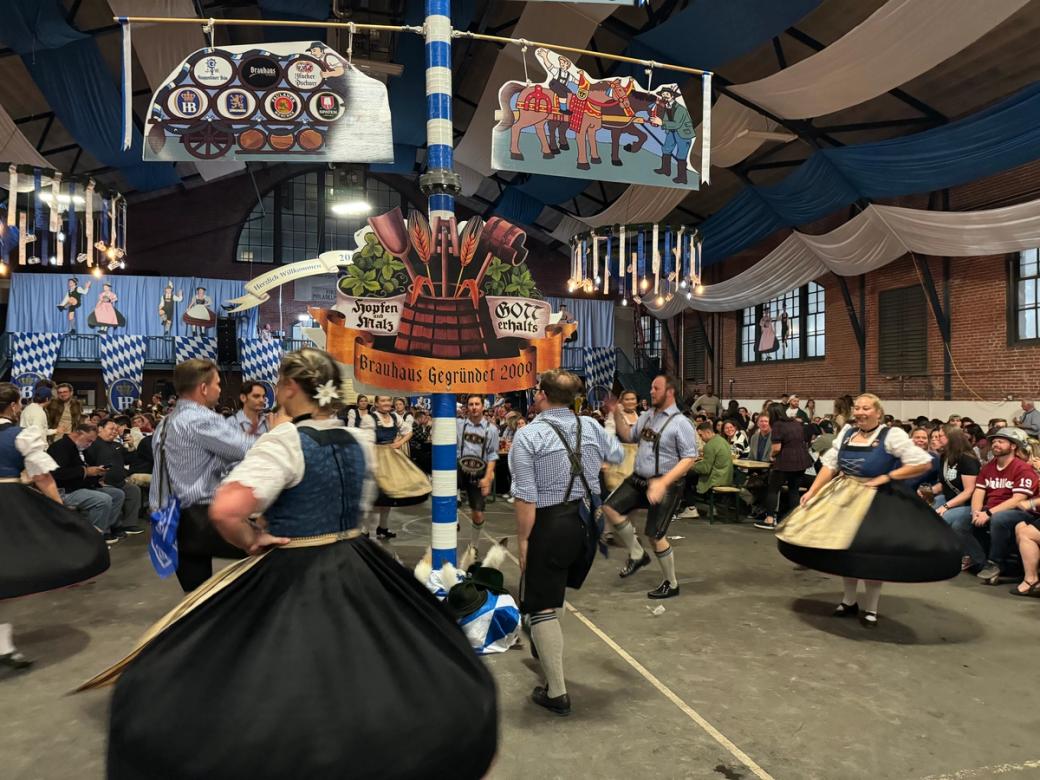  People in traditional German folkwear dance around a pole in front of a crowd. 