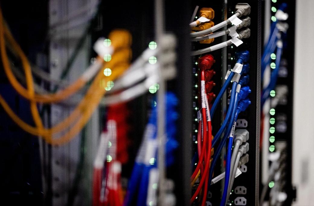A photo of the interior of a server room within a data center with a variety of cords coming out of outlets.