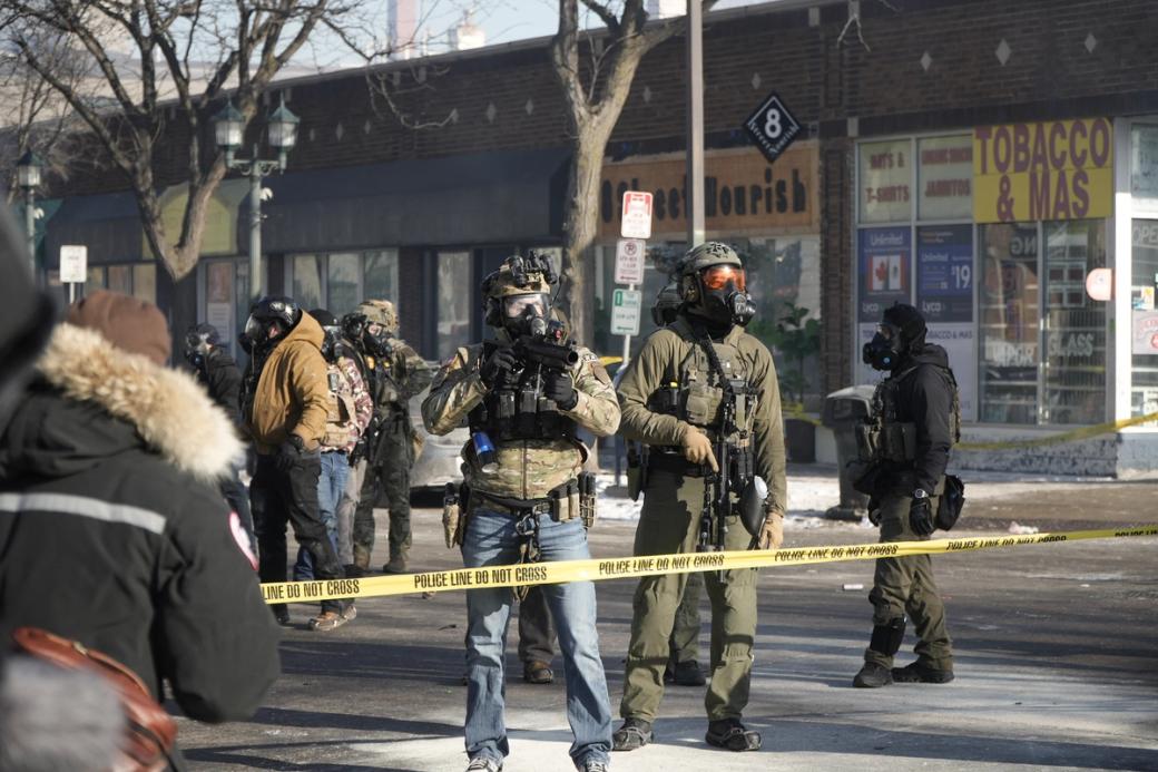 Federal agents in tactical gear and masks hold guns behind police tape