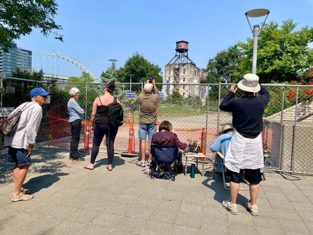 A small group of people gathered in front of a fence blocking access to a heavily damaged building, some trees, and a bridge.