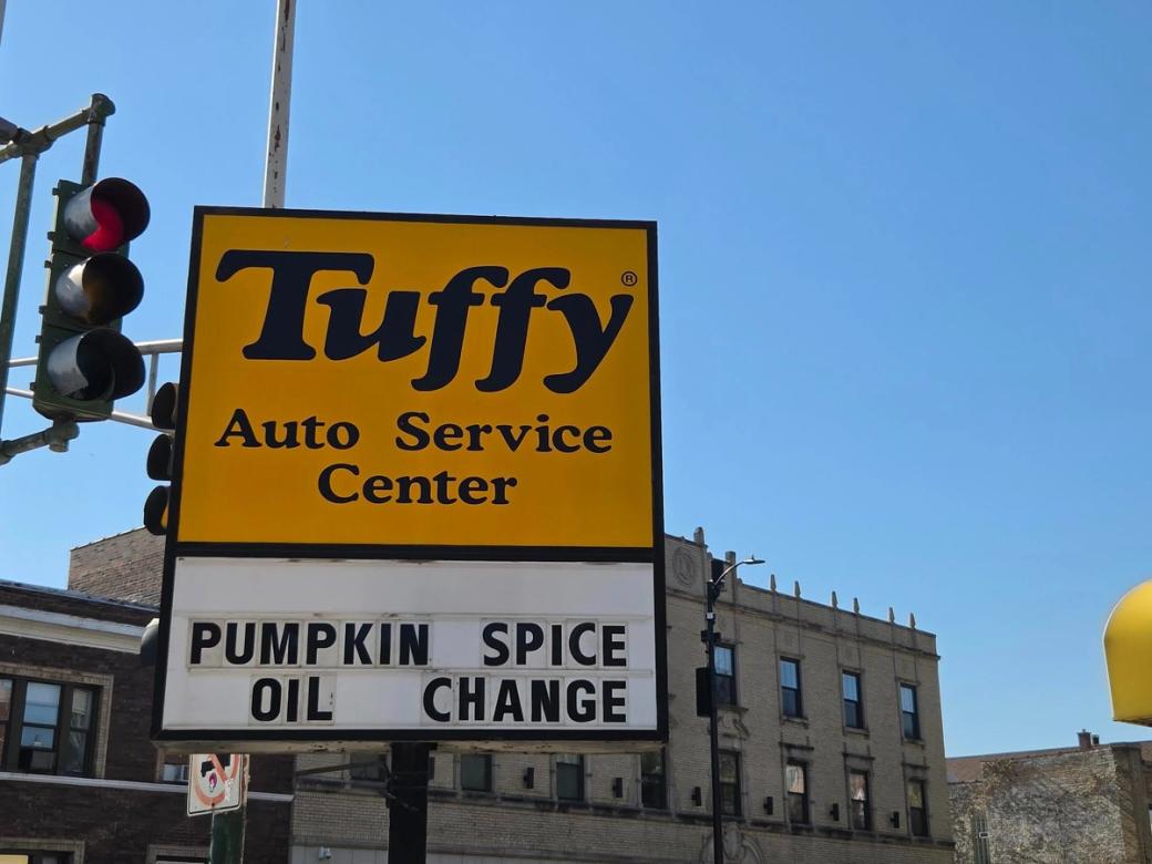 A sunny day with blue skies. There's a yellow sign that says "Tuffy Auto Service Center" below it reads "pumpkin spice oil change"