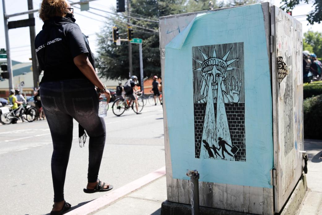 A woman walks by a street poster depicting Lady Liberty refusing an immigrant family to cross a border wall with flood lights for eyes.