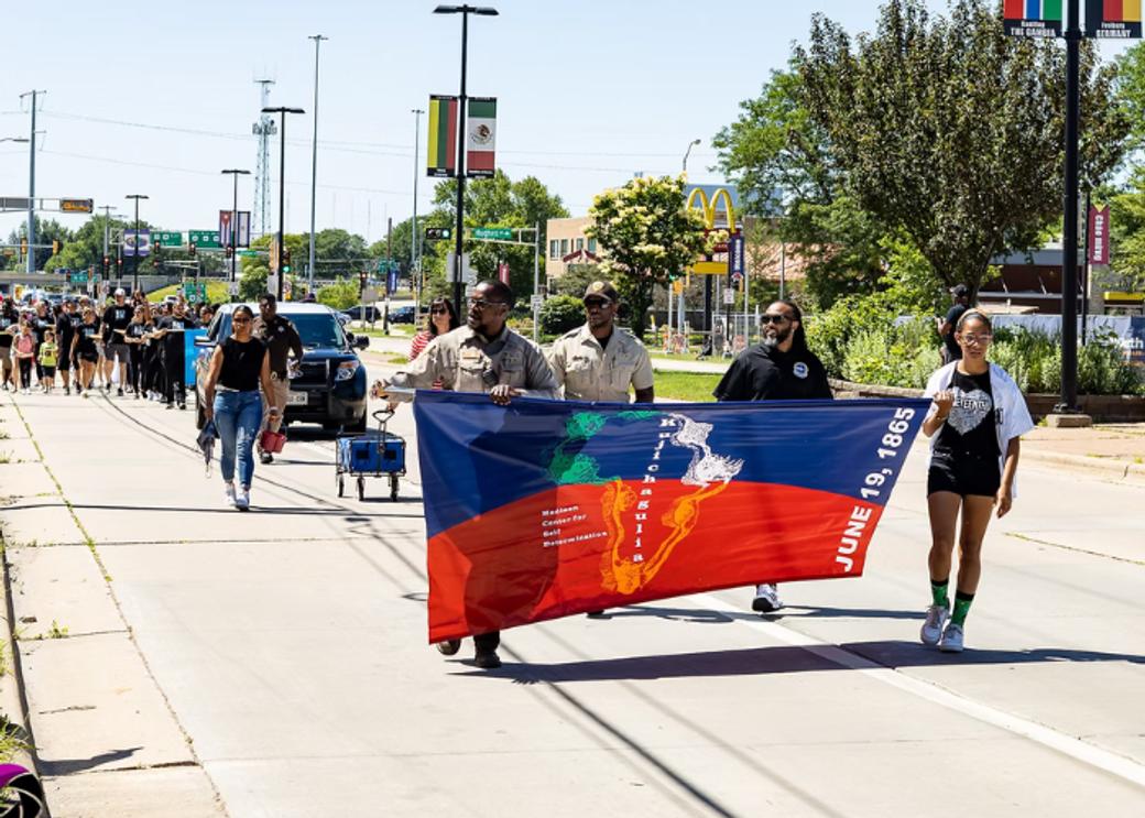 A group of people walk on the street in a parade celebrating Juneteeth. People in the front hold a blue and red banner that reads "June 19, 1865."