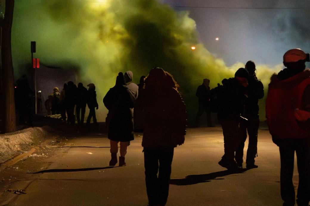 A silhouette of a protester wearing a puffy jacket stands in front of a cloud of green tear gas at night.